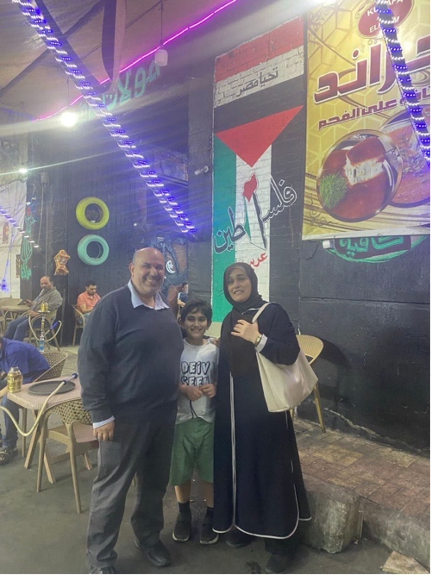 The Nasrallah couple and their child standing on a street corner with a Palestinian flag hung up.