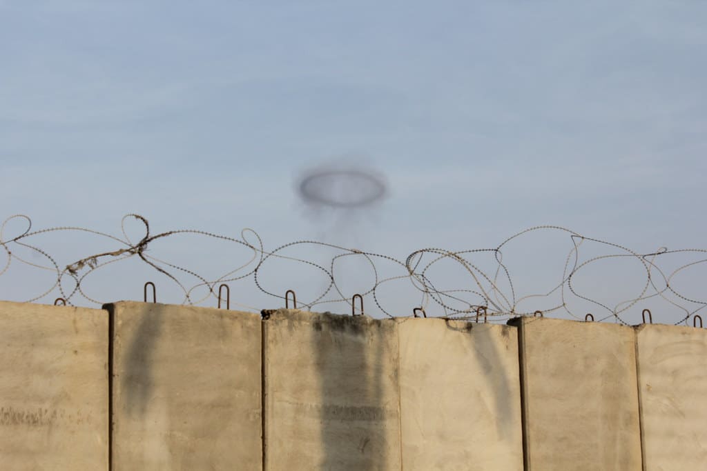 An image of an Israeli bomb hitting Rafah beyond a border wall; the smoke above the wall forms a halo. "Israeli Rockets hitting Rafah, Palestine" by Gigi Ibrahim is licensed under CC BY 2.0.